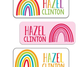 Puede incluir: Tres etiquetas de nombre coloridas con temática de arcoíris con el nombre "HAZEL CLINTON" impreso en cada etiqueta. Las etiquetas son rectangulares y tienen un fondo blanco.