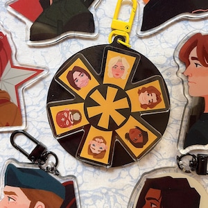 Puede incluir: Un llavero circular negro y amarillo con retratos de personajes. El llavero tiene un cierre amarillo y un anillo negro con un diseño de estrella amarilla en el centro. Rectángulos amarillos con retratos de personajes rodean la estrella.