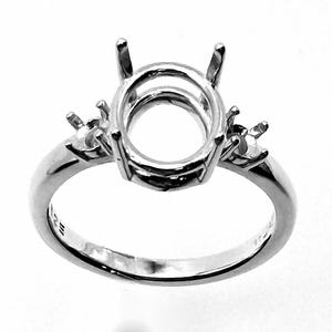 May include: A silver ring setting with an oval center and two side settings. The ring has a polished finish and is designed to hold gemstones. The ring's band is smooth and simple.