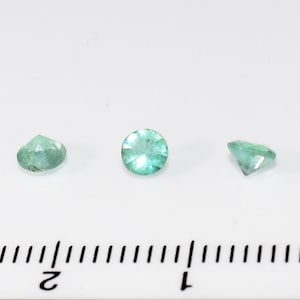 May include: Three round, faceted, light green gemstones on a white surface with a ruler in the background.
