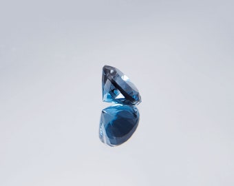 May include: A faceted, oval-cut gemstone in shades of blue, with a dark, almost black, facet. The gem is positioned to show its profile, with its reflection visible below. The background is a gradient of white to light gray.