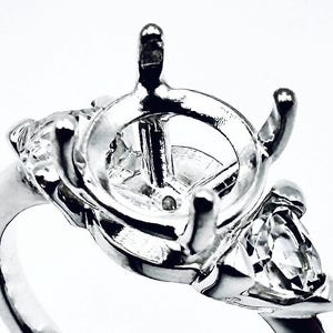 May include: A silver ring setting with a central round mounting and two side settings. The ring features a three-stone design, with the side stones appearing to be pear-shaped. The ring is set against a white background.