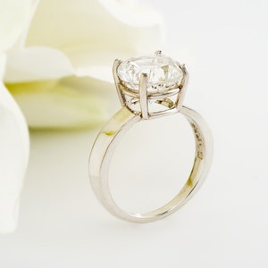 May include: A silver engagement ring with a large, clear, round diamond set in a prong setting. The ring is on a white background.