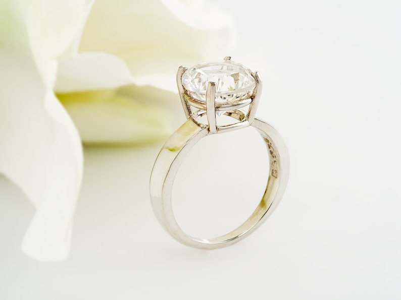 May include: A silver engagement ring with a large, clear, round diamond set in a prong setting. The ring is on a white background.