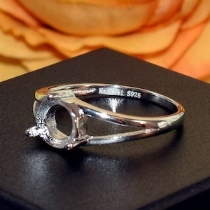May include: A silver ring setting with a split shank design. The ring has a circular setting for a gemstone and is marked with "Mascara L S925". The ring is displayed on a black surface with a blurred floral background.