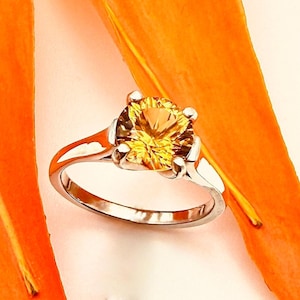 May include: A silver ring with a large, round, yellow gemstone. The gemstone is the focal point, set in a four-prong setting. The ring is displayed against a backdrop of orange and white.