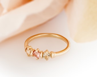 May include: A gold band ring with three small gemstones. The gemstones are a light pink, a darker pink, and a light brown.
