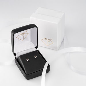 May include: A black jewellery box with a white interior and a small pair of diamond earrings inside. The box is open and the earrings are visible. The box has the word "Mardei" printed on the outside.