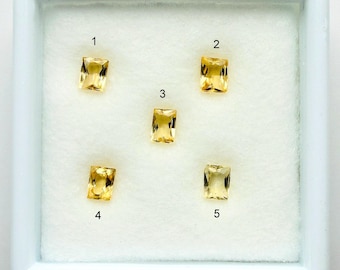 May include: Five rectangular, golden-yellow gemstones are displayed in a white tray. Each gem is numbered from 1 to 5. The gems have a radiant cut, showcasing their clarity and color. The gems are set against a white background.