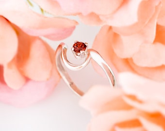 May include: A silver ring with a red gemstone set in a delicate, curved design. The ring is surrounded by soft, peach-colored rose petals.