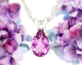 May include: A pear-shaped pendant necklace with a pink gemstone set in a silver setting. The necklace is photographed against a blurred background of purple flowers.