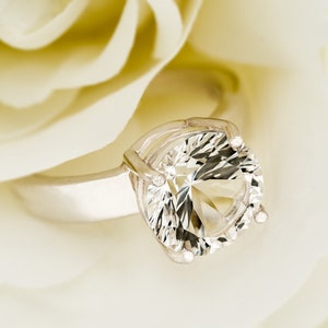 May include: A silver ring with a large, clear, round gemstone set in a prong setting. The ring is resting on a white rose petal.