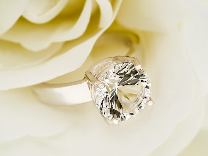 May include: A silver ring with a large, clear, round gemstone set in a prong setting. The ring is resting on a white rose petal.
