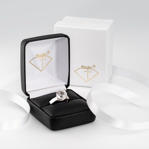 May include: A white ring with a large clear stone in a black ring box with a white interior. The box has the word "Marlei" and a cross inside a diamond shape on the lid. The box is sitting on a white surface with white ribbon.