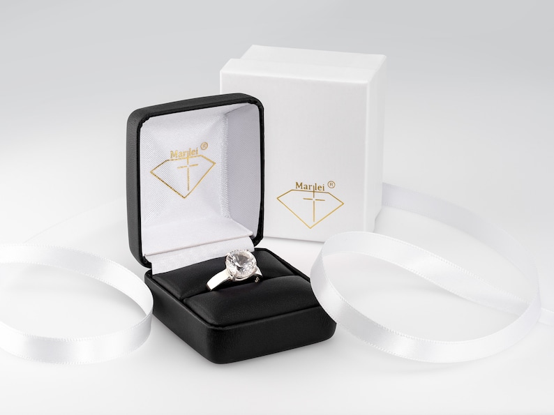 May include: A white ring with a large clear stone in a black ring box with a white interior. The box has the word "Marlei" and a cross inside a diamond shape on the lid. The box is sitting on a white surface with white ribbon.