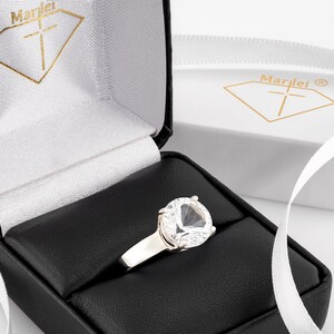 May include: A silver ring with a large, clear, heart-shaped gemstone in a black ring box. The box is lined with white velvet and has the word "Marlei" printed on the inside.