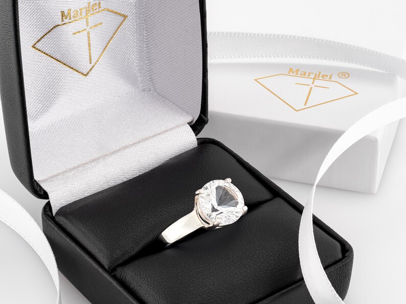 May include: A silver ring with a large, clear, heart-shaped gemstone in a black ring box. The box is lined with white velvet and has the word "Marlei" printed on the inside.