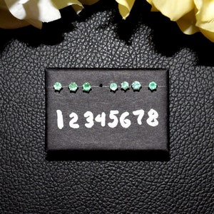 Puede incluir: Una tarjeta negra con ocho espacios numerados, cada uno con una pequeña piedra preciosa verde. La tarjeta está etiquetada con los números del 1 al 8.