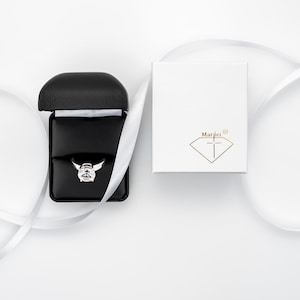 May include: A white ring box with the text "Marlei" and a diamond logo. The box is open and a silver ring with a large clear stone is inside. The box is on a white surface with a white ribbon.