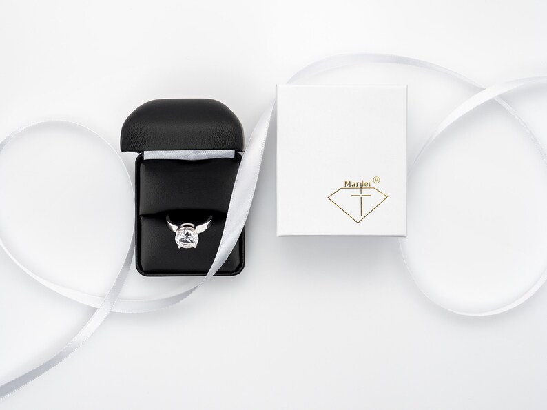 May include: A white ring box with the text "Marlei" and a diamond logo. The box is open and a silver ring with a large clear stone is inside. The box is on a white surface with a white ribbon.