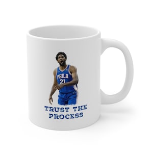May include: White ceramic coffee mug with a basketball player wearing a blue jersey with the word "PHILA" and the number "21" on it. The text "TRUST THE PROCESS" is printed below the player.