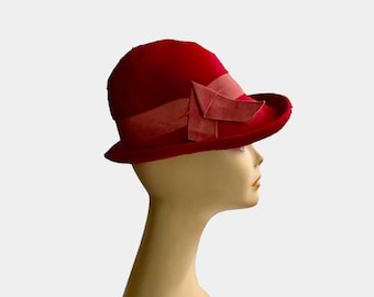 May include: A red cloche hat with a wide brim and a decorative bow. The hat is made of a soft, velvety material and is displayed on a mannequin head. The bow is a lighter shade of red and adds a touch of elegance to the vintage-inspired hat.
