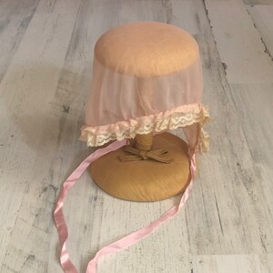 May include: A pink sheer bonnet with a lace trim and pink satin ribbons. The bonnet is displayed on a wooden stand.