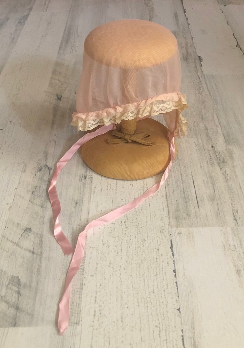 May include: A pink sheer bonnet with a lace trim and pink satin ribbons. The bonnet is displayed on a wooden stand.