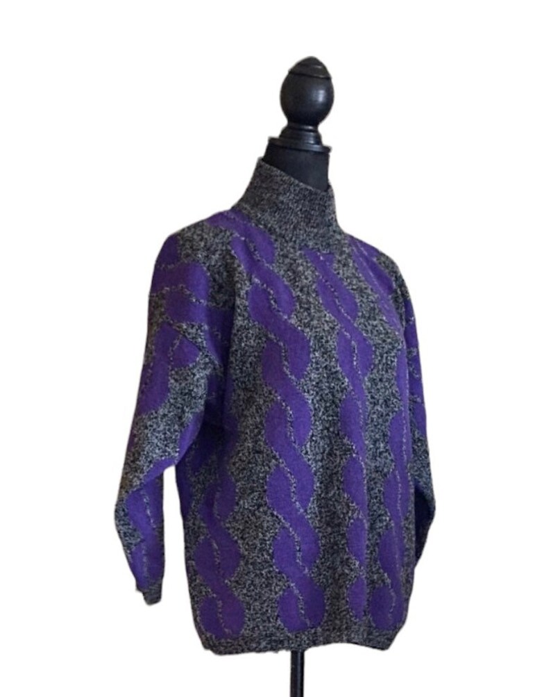 May include: A purple and gray cable knit sweater with a turtleneck. The sweater has a textured pattern of purple cables against a gray background. The long-sleeved sweater is displayed on a black mannequin.