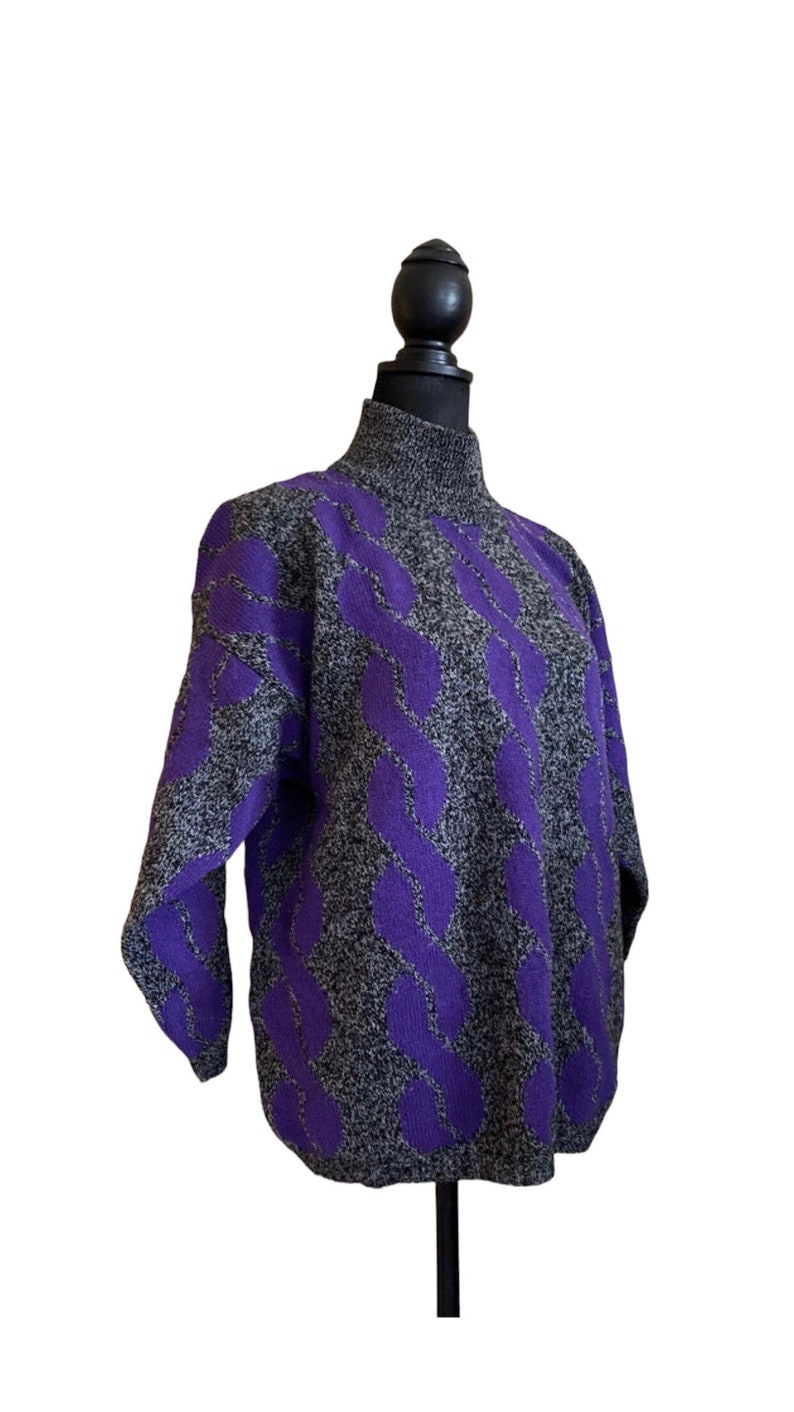 May include: A gray and purple sweater with a high neck and a cable knit design. The sweater is displayed on a black mannequin. The cable knit pattern is a vibrant purple color, and the rest of the sweater is a speckled gray.