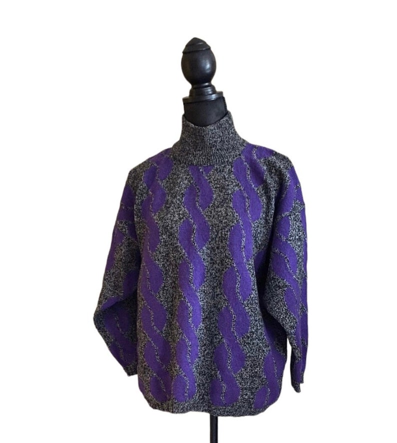 May include: A gray and purple cable knit sweater with a turtleneck. The sweater features a repeating pattern of purple cable knit designs against a gray background. The sweater is displayed on a black mannequin.