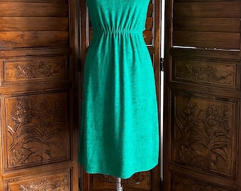 Puede incluir: Un vestido verde brillante hasta la rodilla con escote en V y mangas cortas. La prenda presenta una cintura elástica fruncida y un acabado de tela texturizada, exhibido en un maniquí de costura.