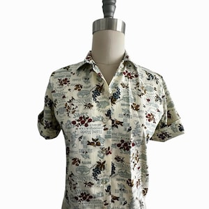 May include: Cream-colored button-down shirt with a repeating pattern of grapes, flowers, and text. The text includes words like "Swiss," "Hand Made," and "Pleasure." The short-sleeved shirt is displayed on a mannequin.