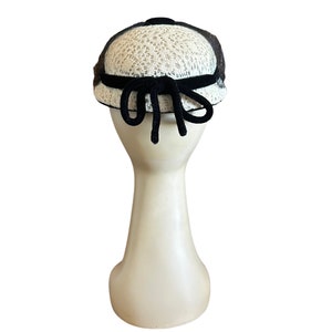 May include: A white and black hat with a black velvet bow. The hat is made of a delicate lace material.