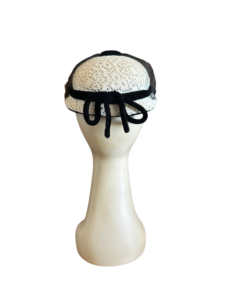 May include: A white and black hat with a black velvet bow. The hat is made of a delicate lace material.