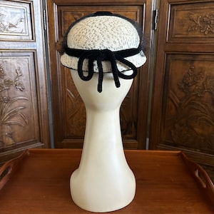 May include: A white and black vintage hat with a black velvet ribbon and bow. The hat is on a white mannequin head.