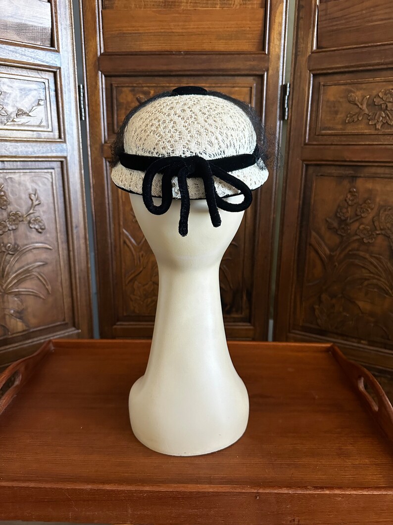 May include: A white and black vintage hat with a black velvet ribbon and bow. The hat is on a white mannequin head.