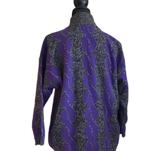 May include: A gray and purple cable knit sweater with a high collar. The sweater features a repeating pattern of purple cable knit designs against a speckled gray background. The sleeves are long and the sweater is displayed on a mannequin.