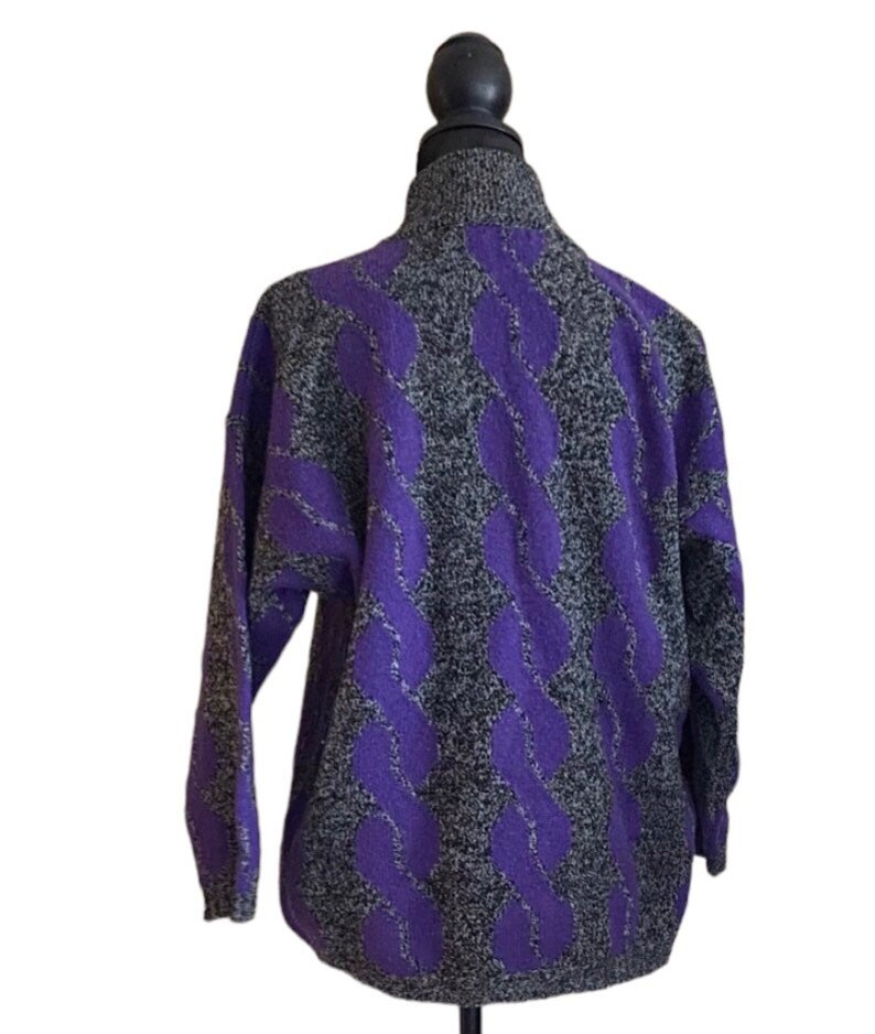 May include: A gray and purple cable knit sweater with a high collar. The sweater features a repeating pattern of purple cable knit designs against a speckled gray background. The sleeves are long and the sweater is displayed on a mannequin.