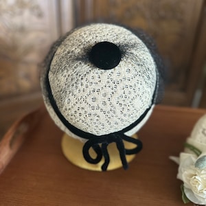 May include: A small, white, lace hat with a black velvet bow and a black velvet button on top. The hat is sitting on a wooden surface.