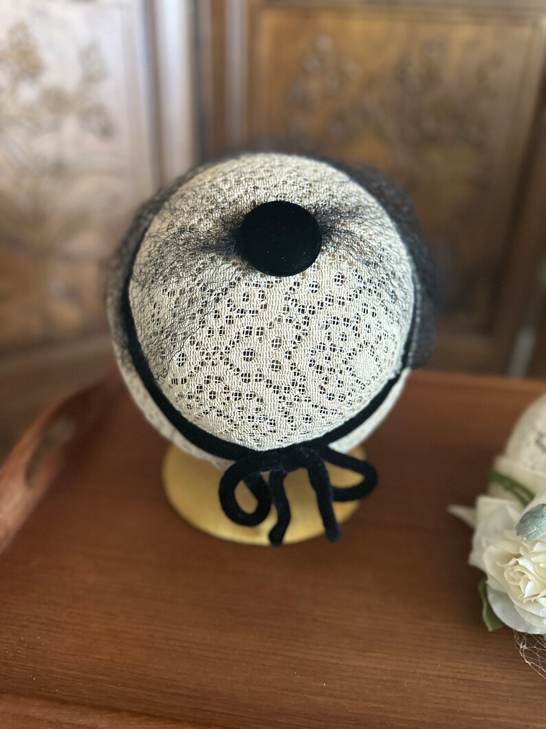 May include: A small, white, lace hat with a black velvet bow and a black velvet button on top. The hat is sitting on a wooden surface.