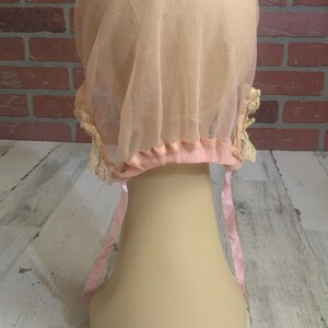 May include: A pale pink tulle bonnet with a satin ribbon tie. The bonnet has a delicate lace trim and is designed to cover the hair.