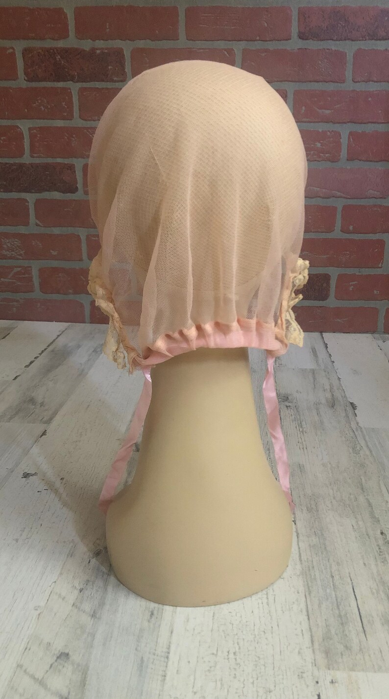 May include: A pale pink tulle bonnet with a satin ribbon tie. The bonnet has a delicate lace trim and is designed to cover the hair.
