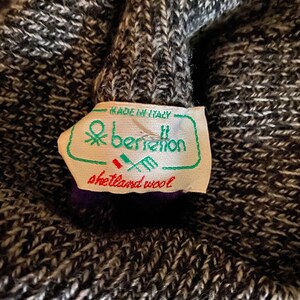May include: Close-up of a gray and white knit fabric with a tag that reads "Made in Italy" and "benetton shetland wool". The tag is green and white with a logo. The knit fabric is textured.
