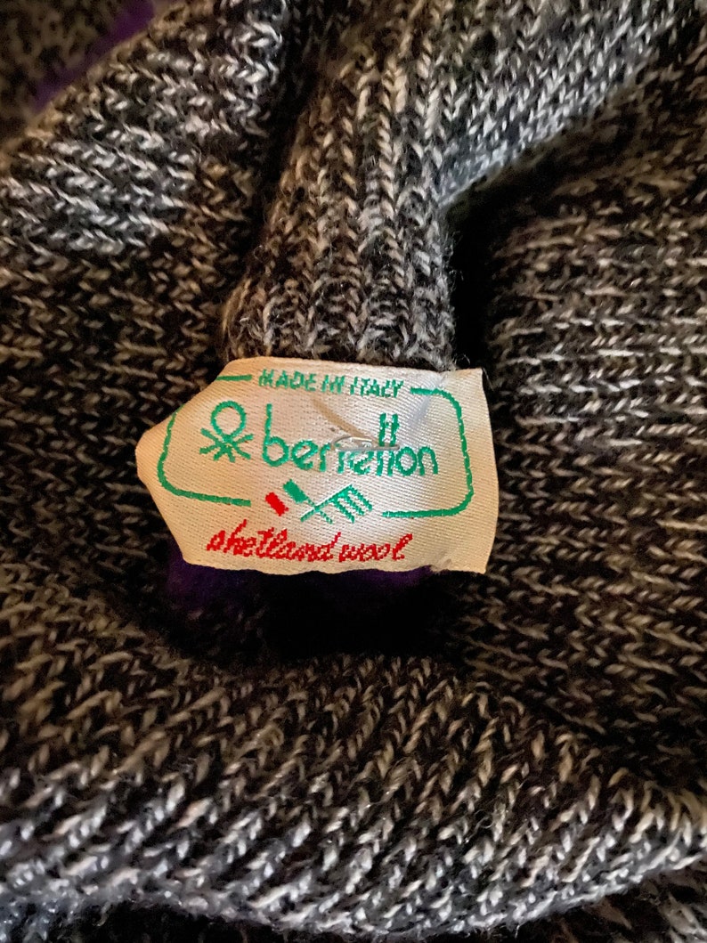 May include: Close-up of a gray and white knit fabric with a tag that reads "Made in Italy" and "benetton shetland wool". The tag is green and white with a logo. The knit fabric is textured.