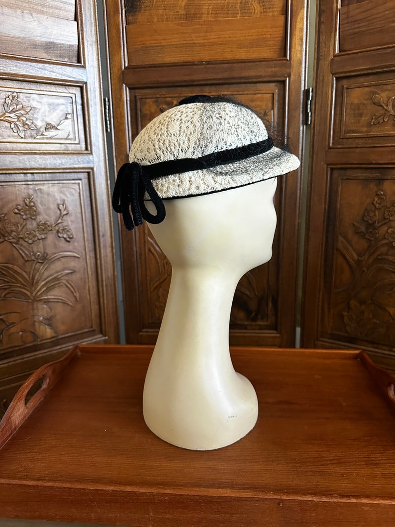 May include: A white and black hat with a black velvet band and a black bow. The hat is on a white mannequin head.