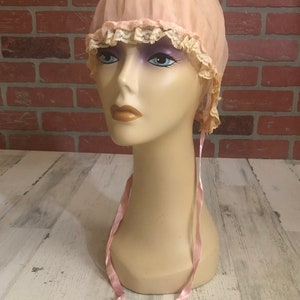 May include: A pink fabric bonnet with lace trim and pink satin ties. The bonnet is on a mannequin head.