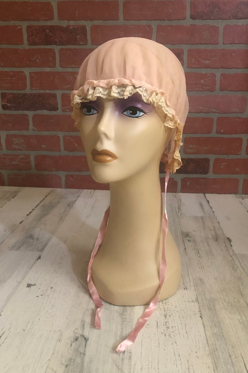 May include: A pink fabric bonnet with lace trim and pink satin ties. The bonnet is on a mannequin head.