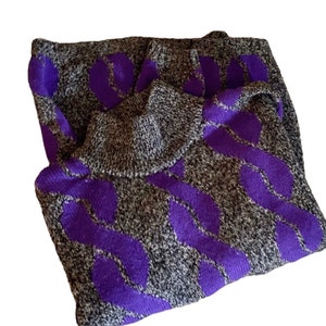 May include: A folded gray sweater with a purple leaf pattern. The sweater has a turtleneck collar and a textured knit design. The purple pattern is a bold contrast against the gray background.