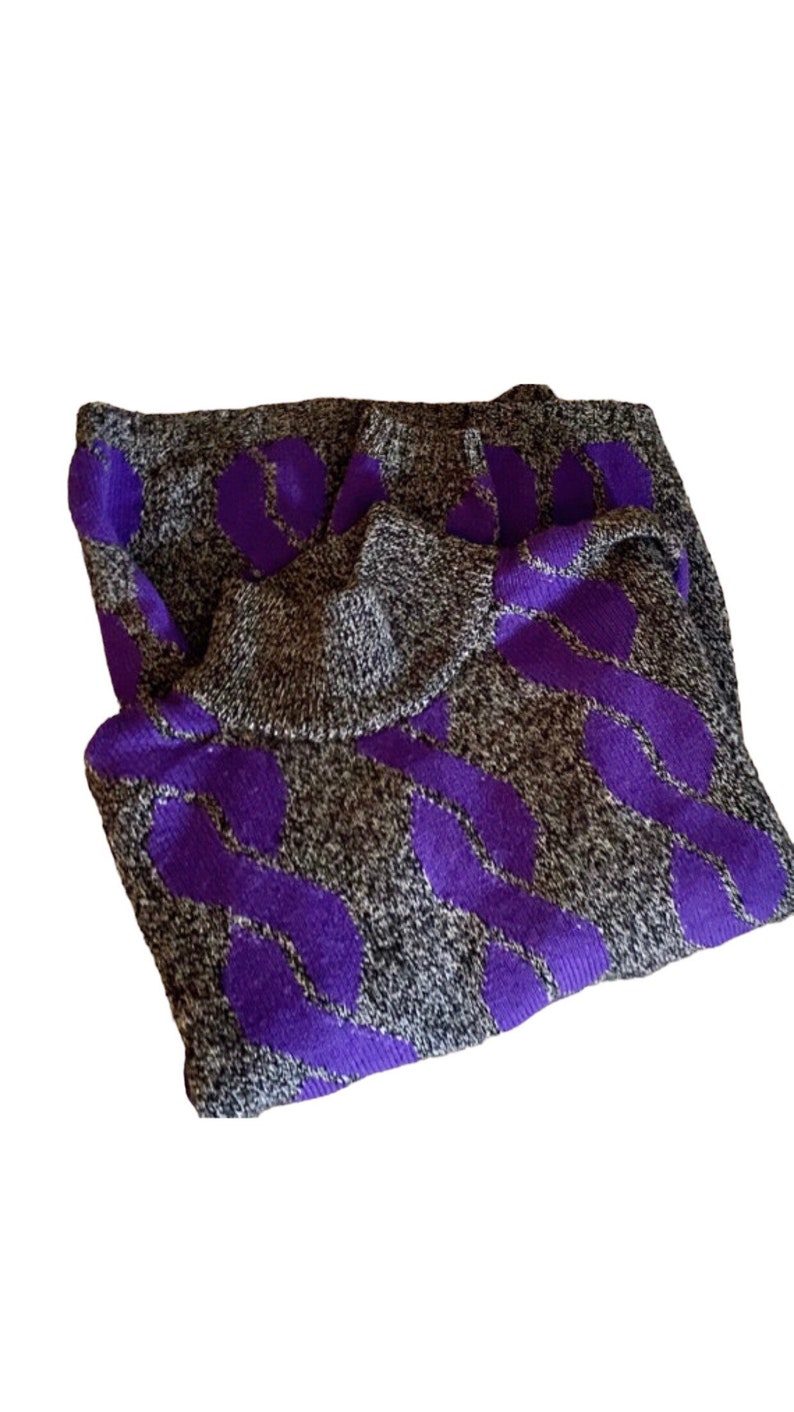 May include: A folded gray sweater with a purple leaf pattern. The sweater has a turtleneck collar and a textured knit design. The purple pattern is a bold contrast against the gray background.